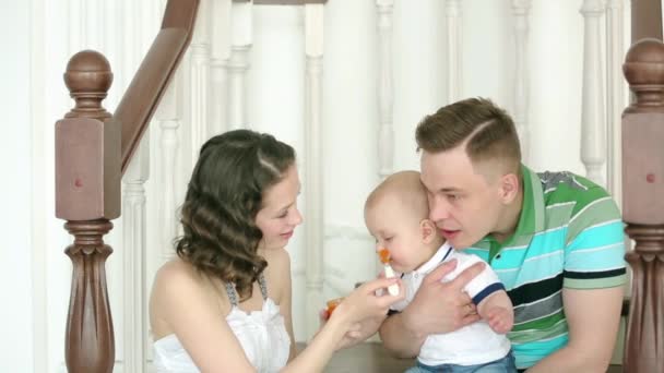 母親が子供をフィードし、お父さんは彼を保持しています。幸せな家族。お母さんお父さんと生後 6 ヶ月の赤ちゃん。家族は家の階段に座って. — ストック動画