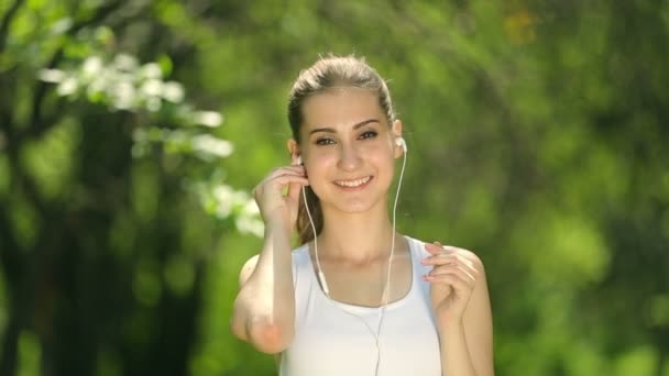 美しい若い女性ランナーの選手は、公園で訓練中に彼女のイヤホンを挿入します。. — ストック動画