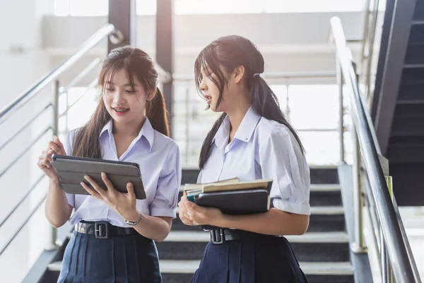 Üniformalı akıllı öğrenci kadınlar modern okul kampüsündeki tabletlerle ev ödevlerini tartışıyor. Taylandlı liseli kız öğrenci.