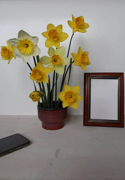 Yellow daffodils stand in a vase and an empty frame on the table.