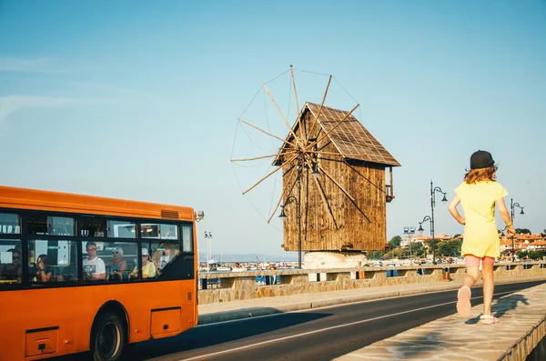 Antiguo molino de viento - uno de los símbolos del casco antiguo de Nessebar. En primer plano, una chica corre por la carretera y los pasajeros del autobús la miran . — Foto de Stock