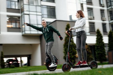 Tek tekerlekli bisiklete binen genç bir adam ve günlük giyinen genç bir kadın şehrin caddesinde elektrikli scooter 'a biniyor.