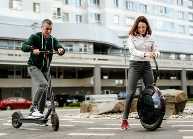 Elektrikli scooter kullanan genç bir adam ve şehir caddesinde tek tekerlekli bisiklete binen günlük giysili genç bir kadın.
