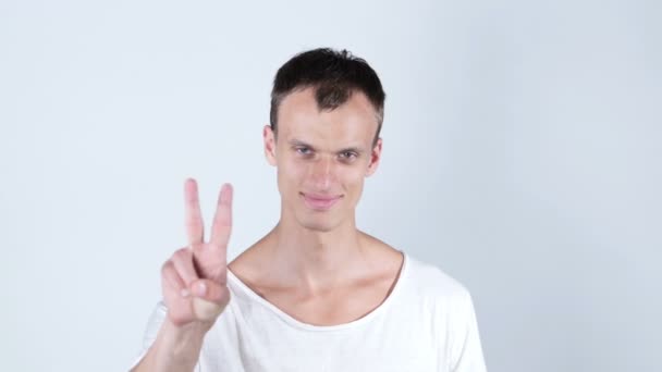 Портрет молодого чоловіка, який посміхається і показує вам знак перемоги на білому тлі — стокове відео