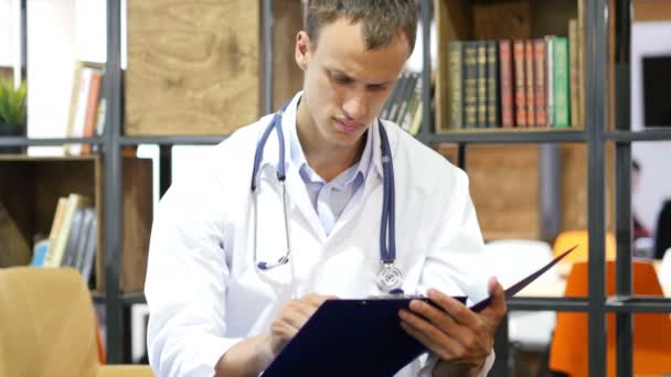 Bonito jovem médico positivo estuda relatório médico em sua clínica — Vídeo de Stock