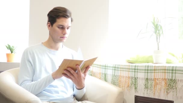 Jovem está sentado em um sofá e lendo um livro — Vídeo de Stock