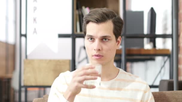 Joven bebiendo agua sentado en un sofá en casa y mirando a la cámara — Vídeos de Stock