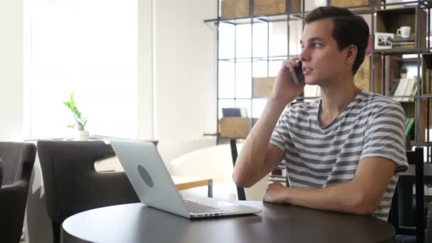 Портрет человека, говорящего по мобильному телефону, сидящего за столом, смотрящего на ноутбук — стоковое видео