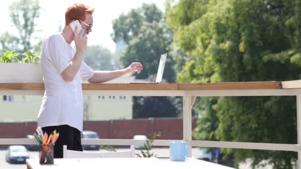 Молодой человек разговаривает по телефону, стоит на балконе на открытом воздухе — стоковое видео