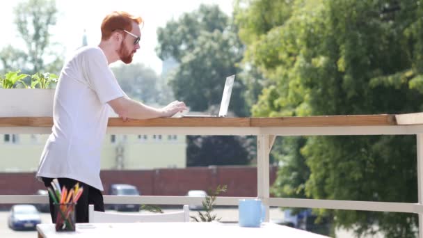 Молодой человек работает на ноутбуке, печатает, стоит на балконе на открытом воздухе — стоковое видео
