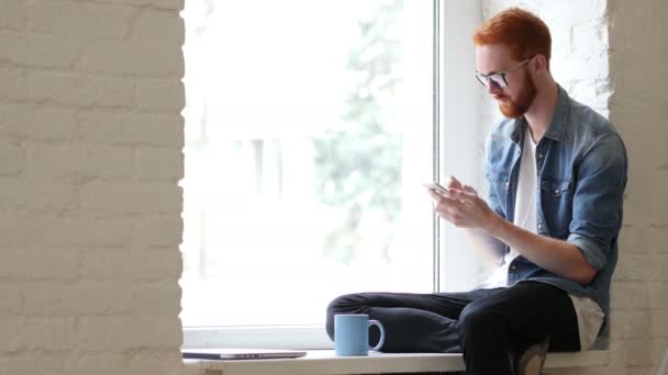 Utilizzando Smartphone, Navigando online da uomo seduto nella finestra, Barba e capelli rossi — Video Stock