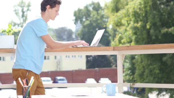 Día soleado, hombre de pie y trabajando en el ordenador portátil en el balcón — Vídeo de stock