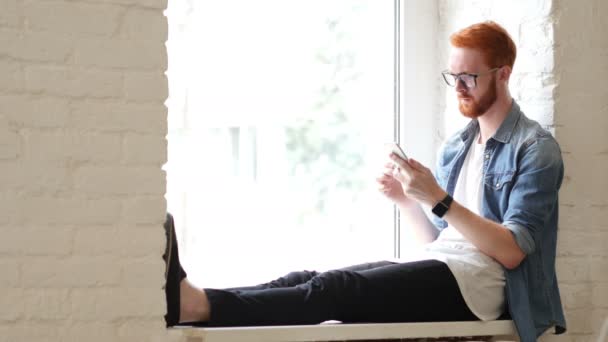 Käyttämällä älypuhelin, istuu ikkunassa, mies w / punaiset hiukset ja parta — kuvapankkivideo