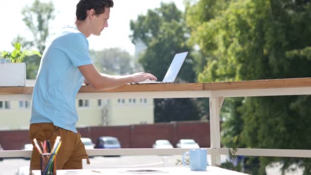 Día soleado, Hombre de pie y mecanografía en el ordenador portátil en el balcón — Vídeo de stock