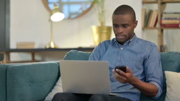 Jonge Afrikaanse man werkt aan smartphone en laptop op de bank — Stockvideo