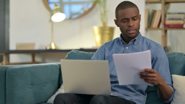 Hombre joven africano con documentos que trabajan en el ordenador portátil en el sofá — Vídeos de Stock