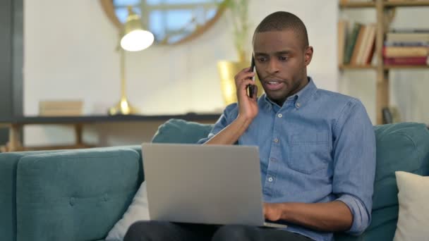 Jonge Afrikaanse Man met Laptop Praten op Smartphone op Sofa — Stockvideo