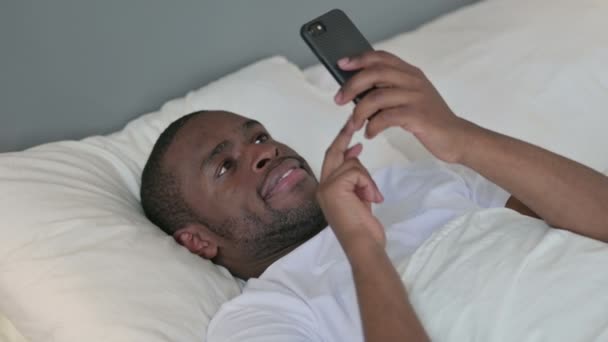 Uso de teléfonos inteligentes por African Man acostado en la cama — Vídeos de Stock