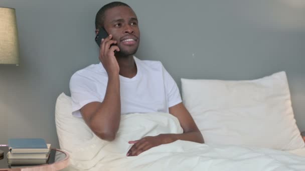 Молодой африканский мужчина разговаривает на смартфоне в постели — стоковое видео