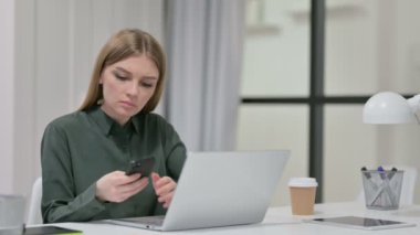 Laptop üzerinde çalışırken akıllı telefon kullanan genç kadın.