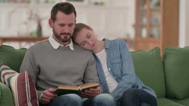 Мужчина читает книгу, пока жена спит на диване — стоковое видео