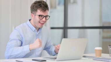 Başarılı genç adam iş yerinde dizüstü bilgisayarında kutlama yapıyor. 