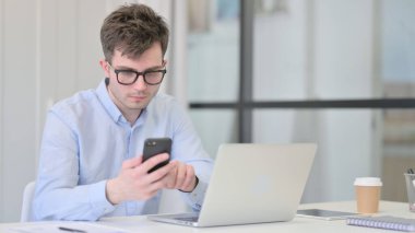 Dizüstü bilgisayarı olan genç adam iş yerinde akıllı telefon kullanıyor.