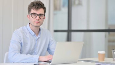 Ofiste dizüstü bilgisayar kullanırken kameraya bakan çekici genç adam.