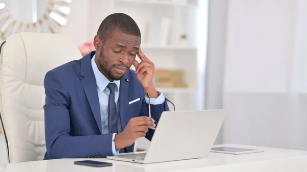 Joven empresario africano con portátil con dolor de cabeza en la oficina —  Fotos de Stock