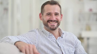 Genç adam koltukta oturuyor ve kameraya gülümsüyor. 