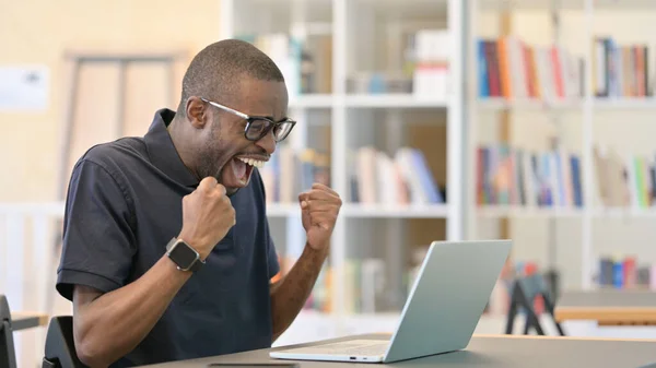 Ambitiös afrikansk man firar framgång på laptop i biblioteket — Stockfoto