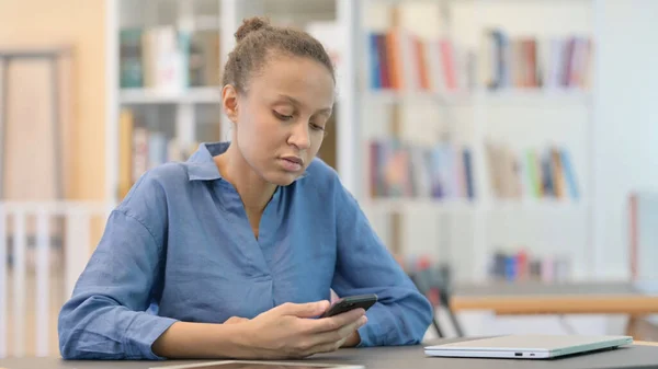 Donna africana annoiata che scorre sullo smartphone in biblioteca — Foto Stock