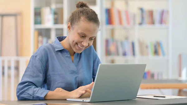 Spännande afrikansk kvinna firar framgång på laptop i biblioteket — Stockfoto