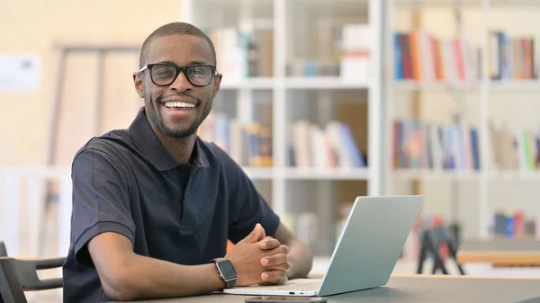 Ung afrikansk man med bärbar dator ler mot kameran i biblioteket — Stockfoto