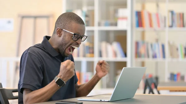 Afrikansk man firar framgång på laptop i biblioteket — Stockfoto