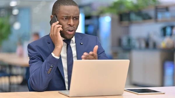 Empresario africano con ordenador portátil enojado en el teléfono inteligente en la oficina —  Fotos de Stock