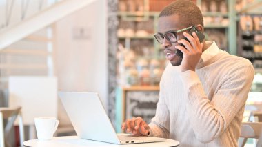 Cafe 'de dizüstü bilgisayarlı Afrikalı adam telefonda konuşuyor.