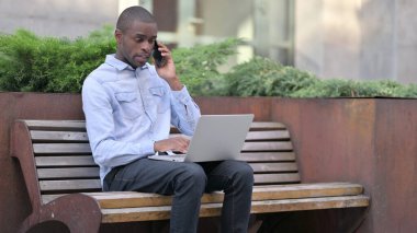 Laptop 'u olan Afrikalı adam dışarıda telefonla konuşuyor. 