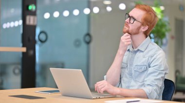 Ofisteki laptopta düşünen ve çalışan dalgın genç kızıl adam. 