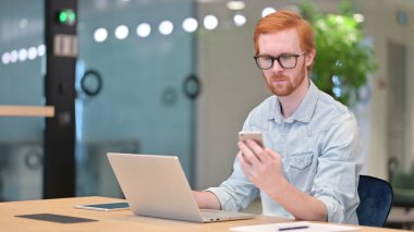 Ofiste akıllı telefon ve dizüstü bilgisayar kullanan kızıl saçlı genç adam. 