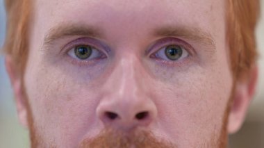 Kızıl saçlı adamın yanıp sönen gözleri kameraya bakıyor. 