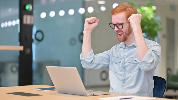 Excited Young Redhead Man Celebrating Success on Laptop in Office
