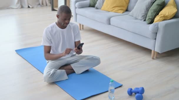 Ung afrikansk man använder smartphone på Yogamattan hemma — Stockvideo