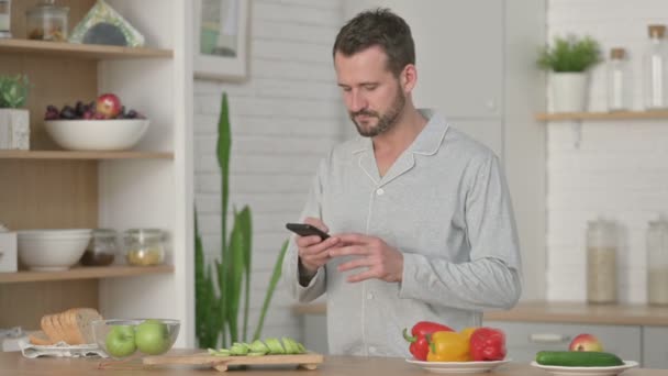 Homem jovem usando Smartphone ao cortar legumes na cozinha — Vídeo de Stock