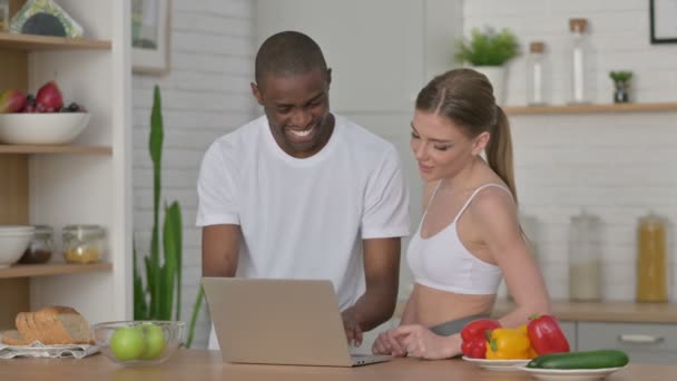 O femeie atletică și un bărbat african lucrează la laptop în bucătărie — Videoclip de stoc