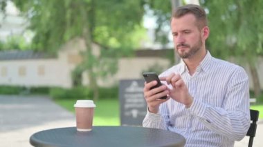 Orta yaşlı adam açık kafedeki akıllı telefondan interneti tarıyor.