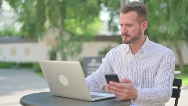 Attraktiver Mann mittleren Alters surft im Outdoor-Café mit Smartphone im Internet — Stockvideo