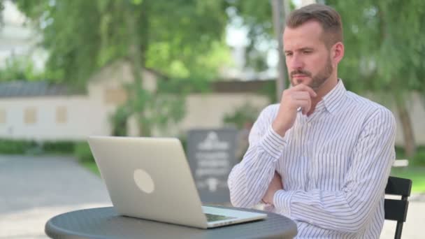 Hombre de mediana edad que piensa mientras trabaja en el ordenador portátil en el café al aire libre — Vídeos de Stock