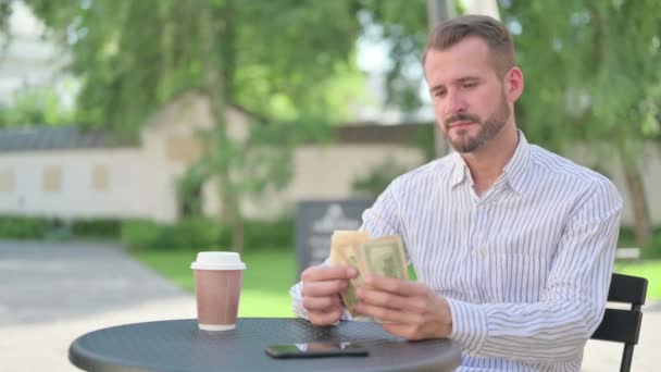 Чоловік середнього віку відчуває себе щасливим під час підрахунку доларів у відкритому кафе — стокове відео