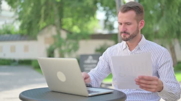 Hombre de mediana edad con documentos de lectura de computadoras portátiles en el café al aire libre — Vídeo de stock